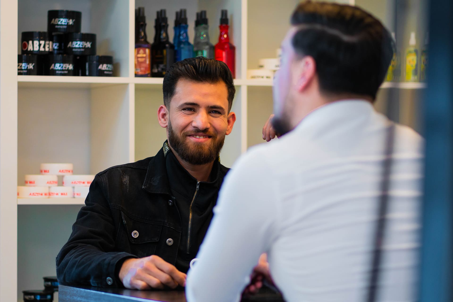 Kapper in gesprek met een klant aan de balie van Kapsalon Assendorp, met haarverzorgingsproducten in de kast erachter.