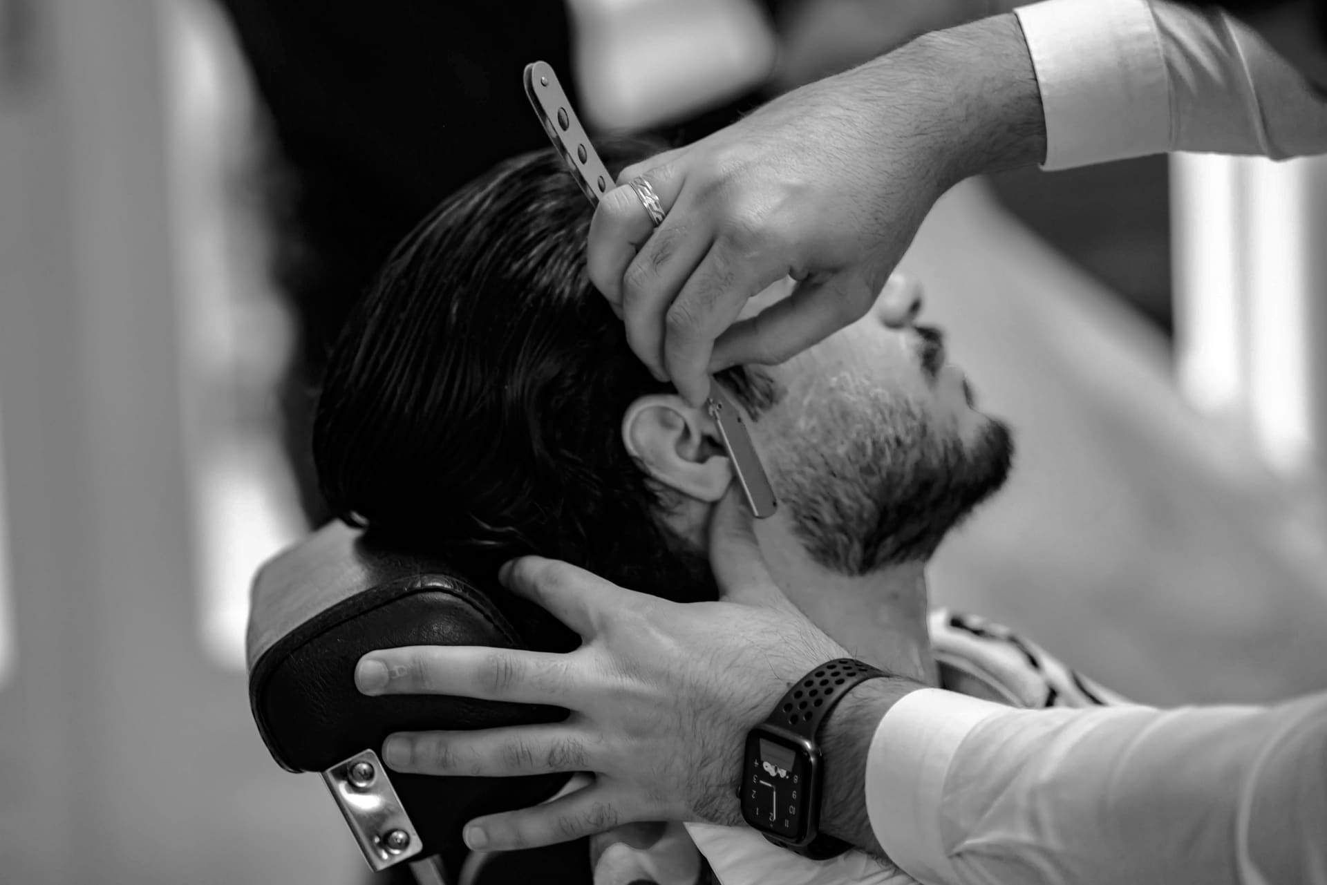 Zwart-witfoto van een kapper die met een scheermes de contouren van een baard bijwerkt.